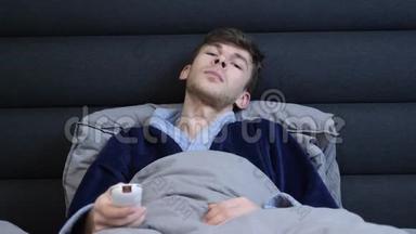 人躺在床上，按遥控器上的按钮。 穿着浴袍的男人躺在床上看电视。 年轻人看电视新闻